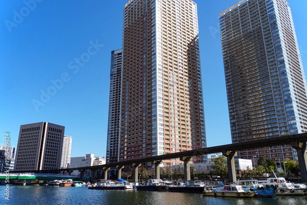 Obraz 晴れ渡った空の下、運河沿いにそびえ立つ複数のタワーマンションとモノレールの高架線