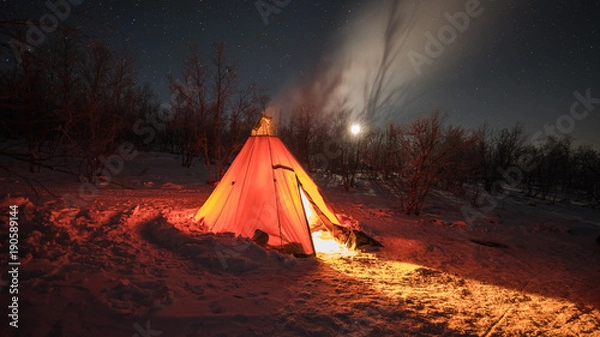 Fototapeta A night photo of arctic landscape with a tee-pee like tent illuminated by a fire inside projecting light out on the snow, Starry clear sky with half moon in the background. (Long exposure time)