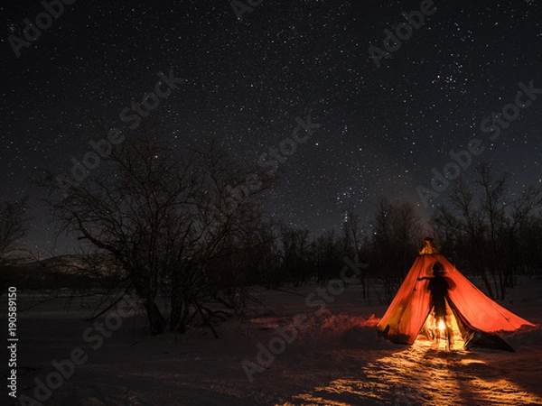 Obraz A night photo of arctic landscape with a tent and a silhouette of a person illuminated by a fire in foreground and starry clear sky in the background. (Long exposure time)