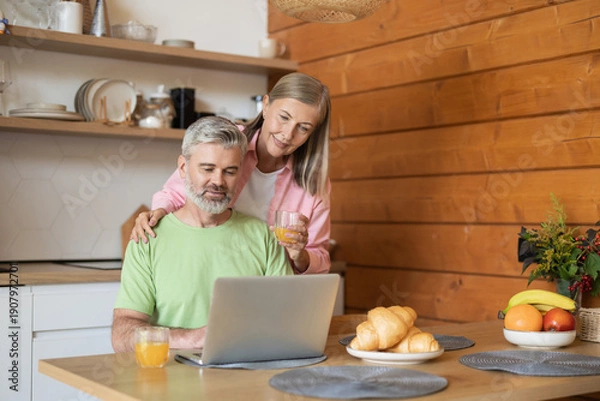 Fototapeta Senior couple using laptop in kitchen while drinking juice