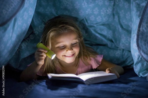 Obraz sweet beautiful and pretty little blond girl 6 to 8 years old under bed covers reading book in the dark at night with torch light smiling happy