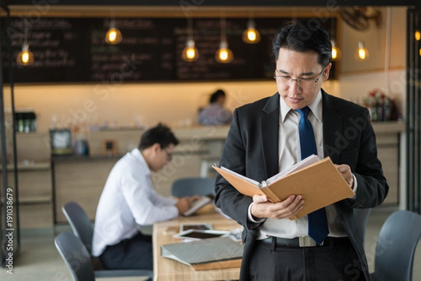 Fototapeta Asian businessman in black suit, who standing and holding document file about financial report on hand.