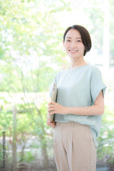 Obraz 綺麗なオフィスの緑背景でオフィスでパソコンを持つミドルの笑顔のカメラ目線の女性　春夏のオフィスカジュアル　縦