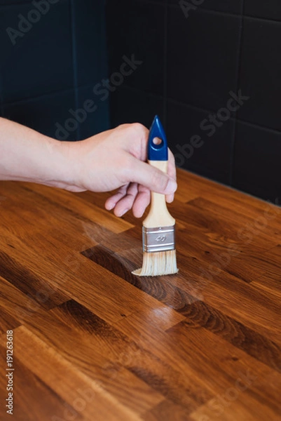 Fototapeta Close up of man's hand applying linseed wood treatment oil with paint brush for protection against water on wooden oak kitchen counter surface after sanding with sanding paper