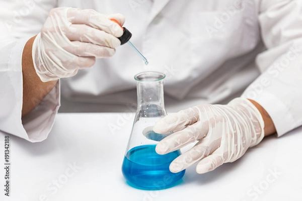 Obraz Hand of a chemist drooping blue liquid chemicals to beaker on the table.