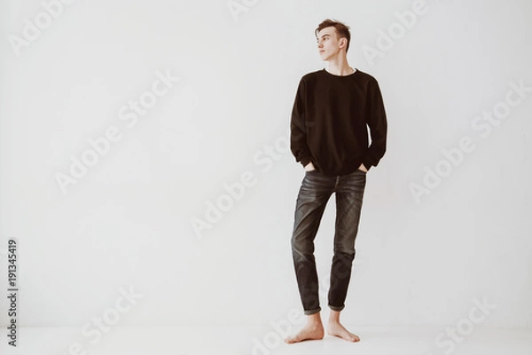 Fototapeta A young man, a student stands against a white wall in the room, dressed in short jeans and a black sweater, barefoot. The guy is a model.