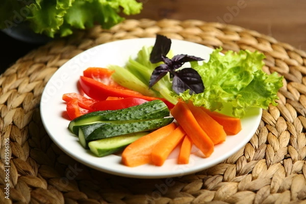 Obraz Pepper, carrots, celery, raw cucumber are cut into strips. On the plate. Next to it are lettuce leaves, basil. On a background a wicker stand, a wooden table, a black background.