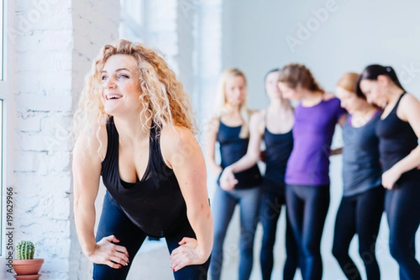 Fototapeta Close up image of attractive curly blond fitness woman over blurred group of people in white gym interior. Teamwork, good mood and healthy lifestyle concept.