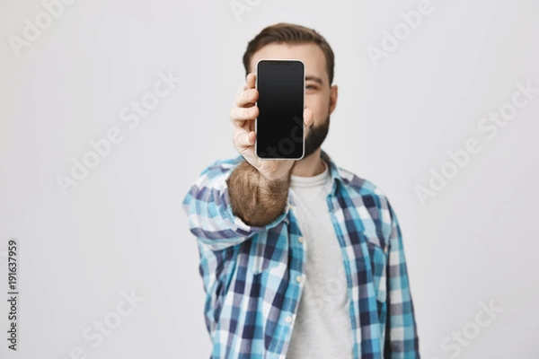 Fototapeta Male model with beard and trendy haircut in checked shirt showing smartphone at camera covering face with it, standing against gray background. Consultant shows gadget to customer of shop he works at