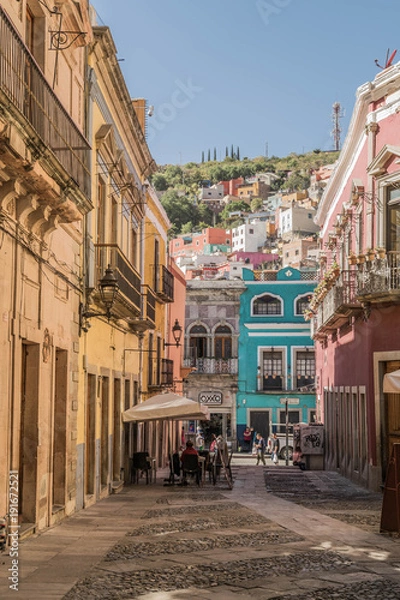 Fototapeta A charming cobblestone pedestrian street with colorful buildings, outdoor table, and a hill dotted with colorful homes, in the background, in Guanajuato, Mexico