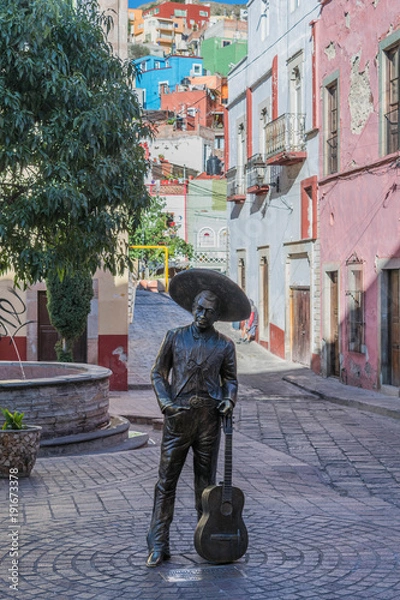 Fototapeta The statue in the Plaza del Ropero, with decorative stone pavement, partial view of a tree, and a narrow street heading up to colorful houses dotting the hillside, in Guanajuato, Mexico