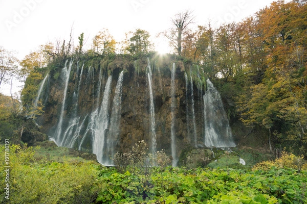 Obraz The sun shining above a waterfall in Plitvice Lakes National Park. The are lush green plants in the foreground and orange and yellow trees in the background above the waterfall. 