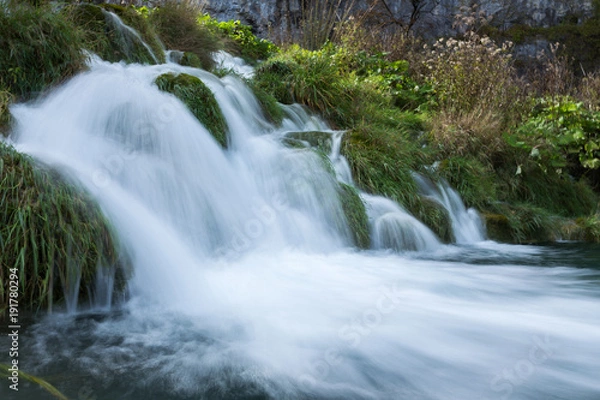 Obraz A long exposure close up at the bottom of a small waterfall. The grass is soaking wet and with the sun shining on part of it. 