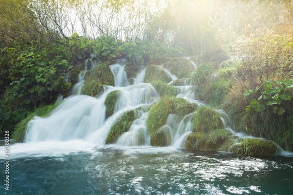 Obraz A sunny day looking at a small waterfall surrounded by lush green grass and turquoise waters. The background is a golden yellow. This is a long exposure.