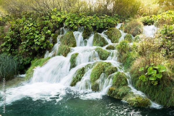 Obraz Light leak from the sun over a small waterfall in Plitvice Lakes national Park. The water is flowing in different directions and it passes the lush green grasses. The water is turquoise.
