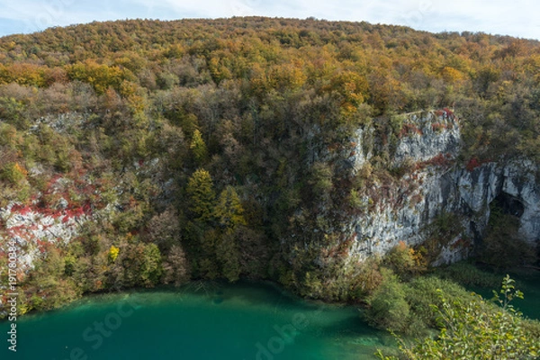 Obraz A gigantic white cliff covered in colorful trees in Plitvice Lakes National Park. The water is a turquoise green with clouds above.