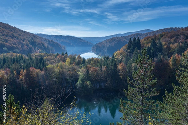 Obraz Two lakes are shown in Plitvice Lakes National Park. The trees are changing colors from green to yellow, orange and red. There is some fog above the lake and few clouds in the blue sky. Great distance