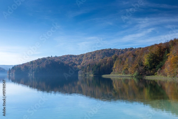 Obraz Reflection of a peninsula on a lake. Taken in Plitvice Lakes National Park. The sky is mostly clear with few clouds and the trees are turning colors.