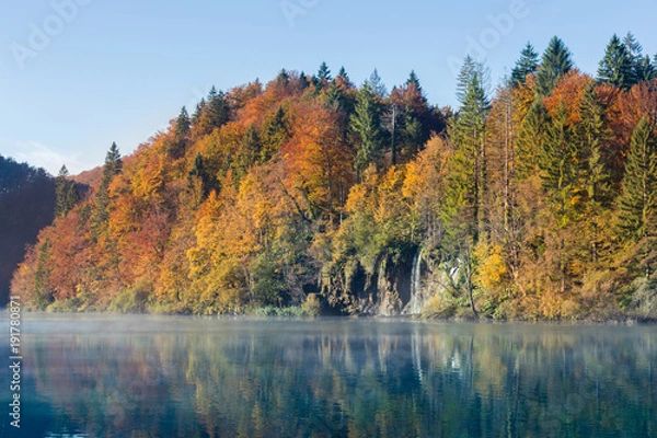 Obraz A waterfall flows into a turquoise lake in autumn. The trees are red orange green yellow and the sky is clear. There is a layer of mist or fog on top of the still water.