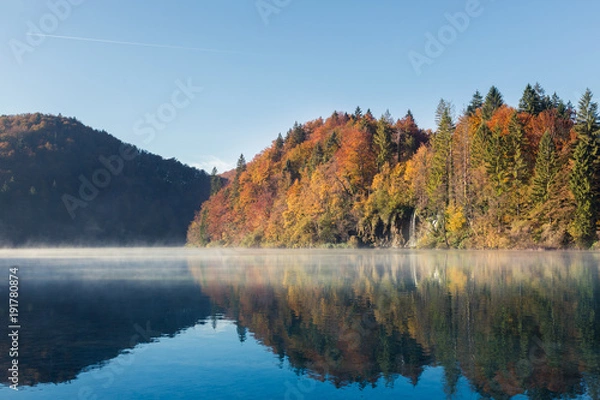 Obraz Early morning on a calm lake in Plitvice Lakes National Park. In the background there is a waterfall flowing into the lake. The trees are colored red orange green and yellow. There is a layer of fog.