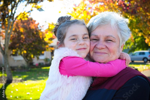 Obraz Happy family, generation and people concept - happy smiling young tree years old granddaughter with senior grandmother standing by House at Autumn Season, hugging in Oregon USA close-up portrait 