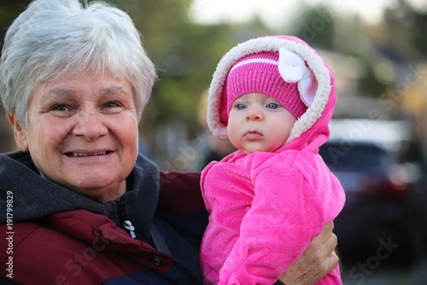 Obraz Happy family, generation and people concept - happy smiling young tree years old granddaughter with senior grandmother standing by House at Autumn Season, hugging in Oregon USA close-up portrait 