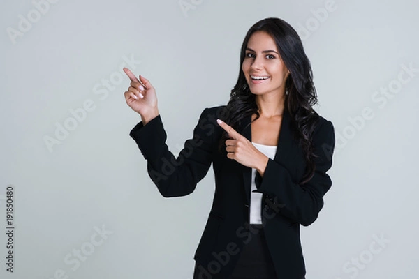 Fototapeta Look here! Beautiful young businesswoman pointing away and looking at camera with smile while standing against white background