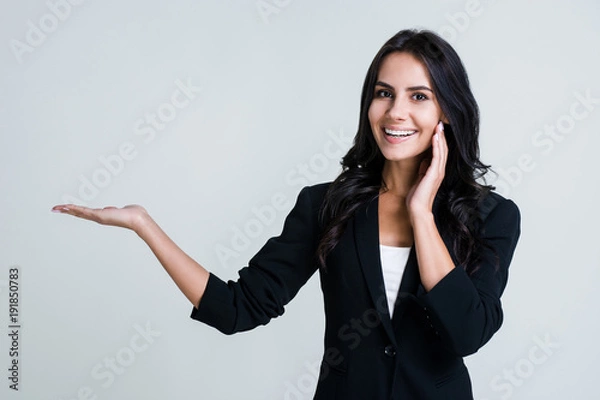 Fototapeta Have you seen this? Beautiful young businesswoman pointing away and looking at camera with smile while standing against white background