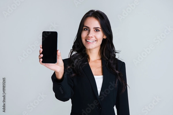 Fototapeta Have you seen this app? Beautiful young businesswoman showing her smartphone and looking at camera with smile while standing against white background