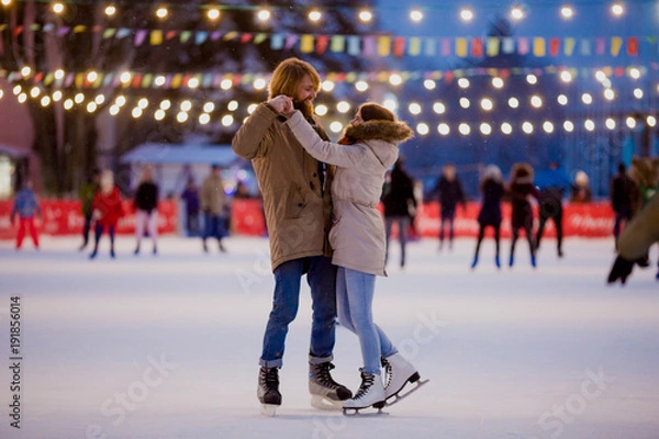 Fototapeta Theme ice skating rink and loving couple. meeting young, stylish people ride by hand in crowd on city skating rink lit by light bulbs and lights. Ice skating in winter for Christmas on ice arena