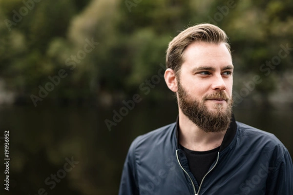 Fototapeta One bearded man wearing a blue jacket, standing in front of a lake and trees on a moody day.