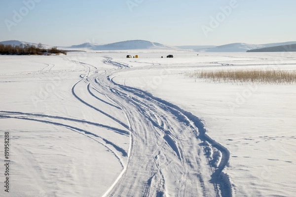 Fototapeta Trace from cars on a snowy lake with fishermen in the background