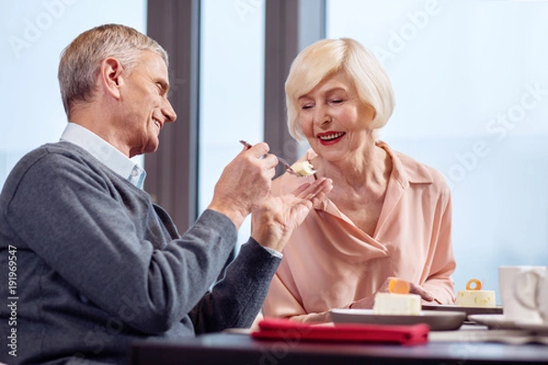 Obraz Sweet date. Handsome cheerful mature man sharing desert with wife while smiling and having date