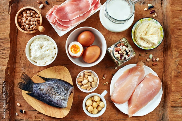 Fototapeta Flat lay of high-protein foods on rustic wooden table. Milk, eggs, cottage cheese, fish, chicken, pork, chickpea, beans and nuts.