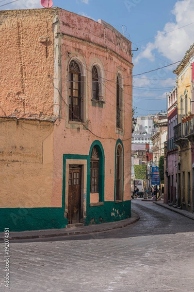 Fototapeta A street scene with a curve corner building painted yellow and green, a cobblestone street and other architectural details, in Guanajuato, Mexico