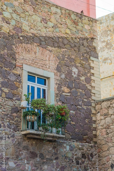 Fototapeta Aged stone wall with a long window, a balcony with some potted plants, and a sliver of pink, in the background, in the old city of Guanajuato, Mexico-a UNESCO Heritage site
