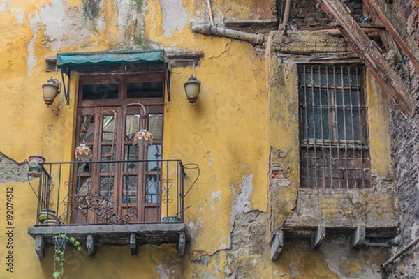 Fototapeta Old and decaying outer city wall, with a balcony with french wooden doors and iron railing, a double wooden window with iron bars, and yellow chipping paint, in Guanajuato, Mexico