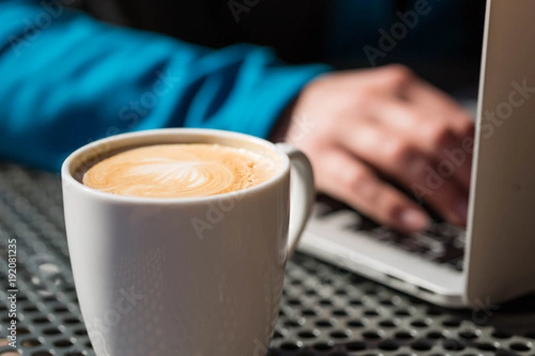 Fototapeta Coffee latte on a table from a cafe, with foam while a person works on a laptop computer