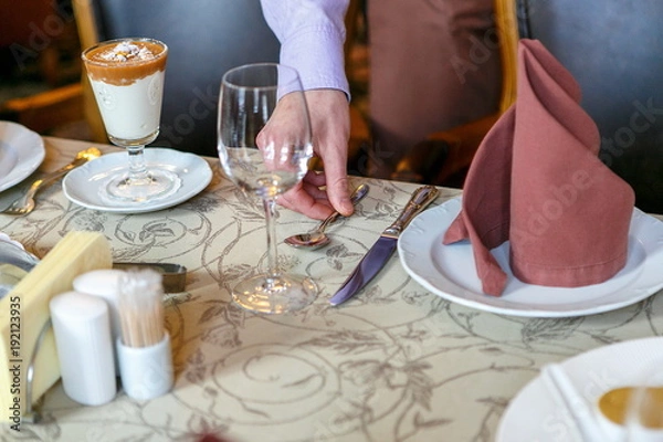 Obraz Waiters prepare a table for breakfast. Background. The foreground of napkins, dishes, glasses. Calm atmosphere. Close up. Hand adjusts the spoon