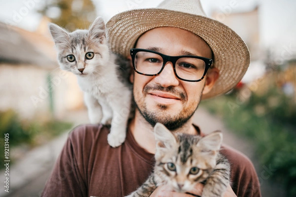 Obraz Happpy young bearded farmer holding two little kitten in hands outdoor in village with abstract background. Smiling man in glasses and straw hat playing with funny cute pets. Have fun in countryside.