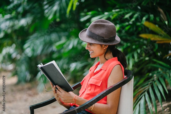 Fototapeta Portrait of a young and attractive Indian Asian girl reading a book in the sun during the day while sitting in a chair in a green park. She is smiling and enjoying herself and is fashionably dressed.
