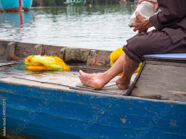 Fototapeta Vietnamese fisherman in traditional clothes closeup sitting in a wooden boat without a foot on the left leg