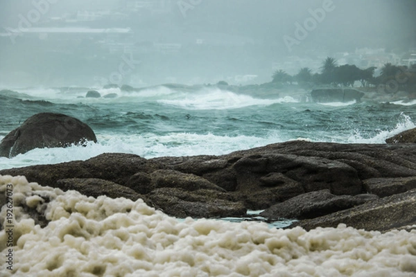 Fototapeta Crashing waves onto rocks on a misty day with Camps Bay, Cape Town in the background.
