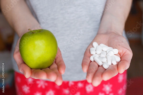 Fototapeta Woman holds a green apple in one hand and a handful of pills in the other.