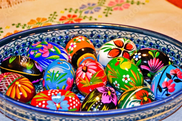 Fototapeta Colorful Hand Painted Wooden Easter Eggs in a Ceramic Bowl on a cloth with a needlepoint flower design.