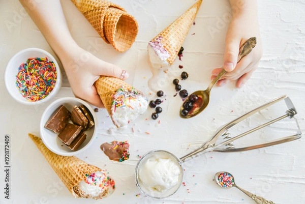 Obraz The child Makes his own Ice cream: Top View, Children's Hands Holding a cone of ice Cream On the table Chocolate, Spoon, Topping, Berries, Sprinkles, Jam.