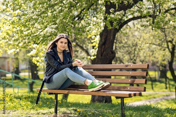Fototapeta Glamorous young Caucasian woman in black leather jacket sitting in the park on the bench