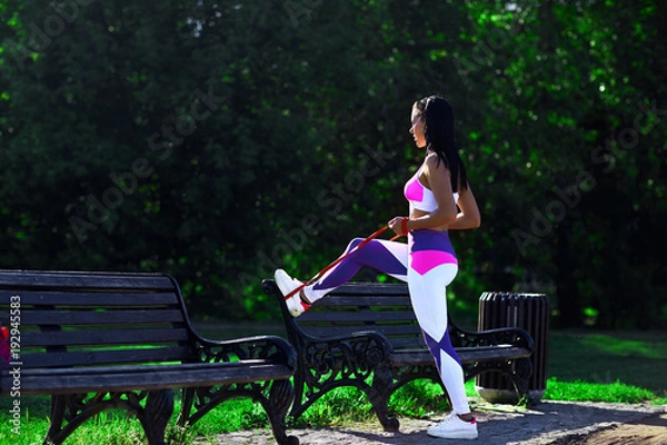 Obraz Sporty woman with dark hair does exercises with the expander on a Park bench.