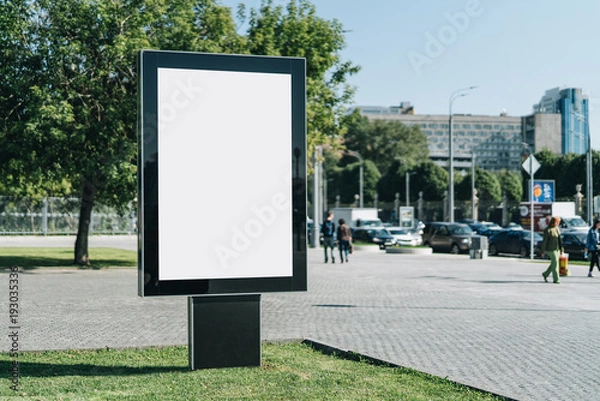 Fototapeta Vertical blank glowing billboard on the city street. In the background buildings and road with cars. Mock up. The poster on the street next to the roadway.