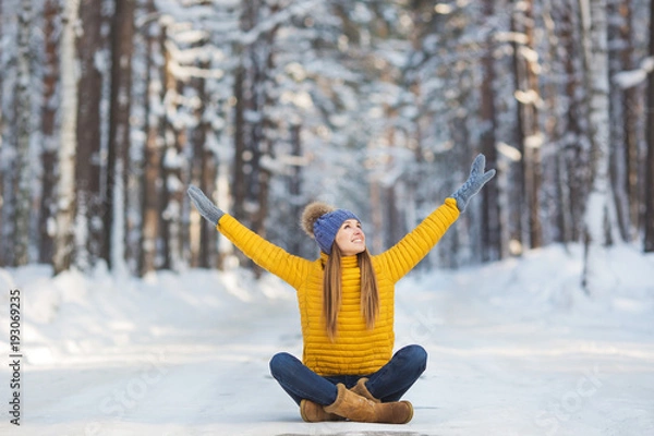 Fototapeta Young smiling woman in a bright clothes sits on a road and hands up in a winter forest